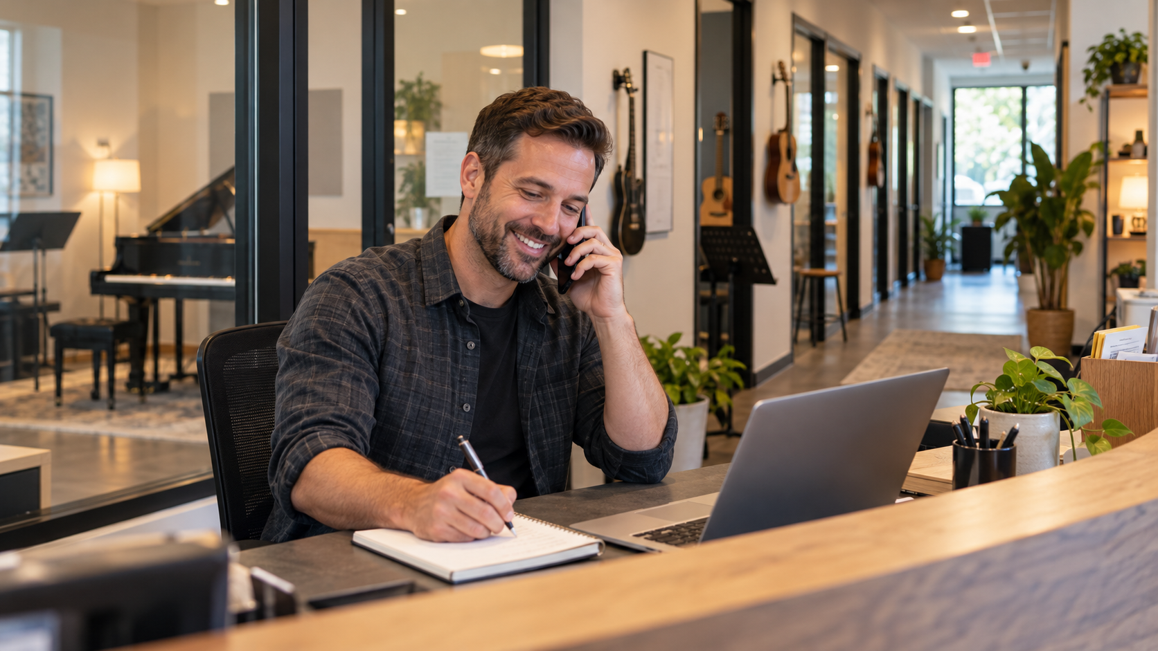 A smiling man on the phone at a music school front desk, taking notes beside an open laptop, with a grand piano, guitars on the wall, and lesson rooms down the hall behind him.